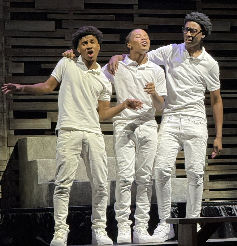 Three young actors in white singing with arms around each other